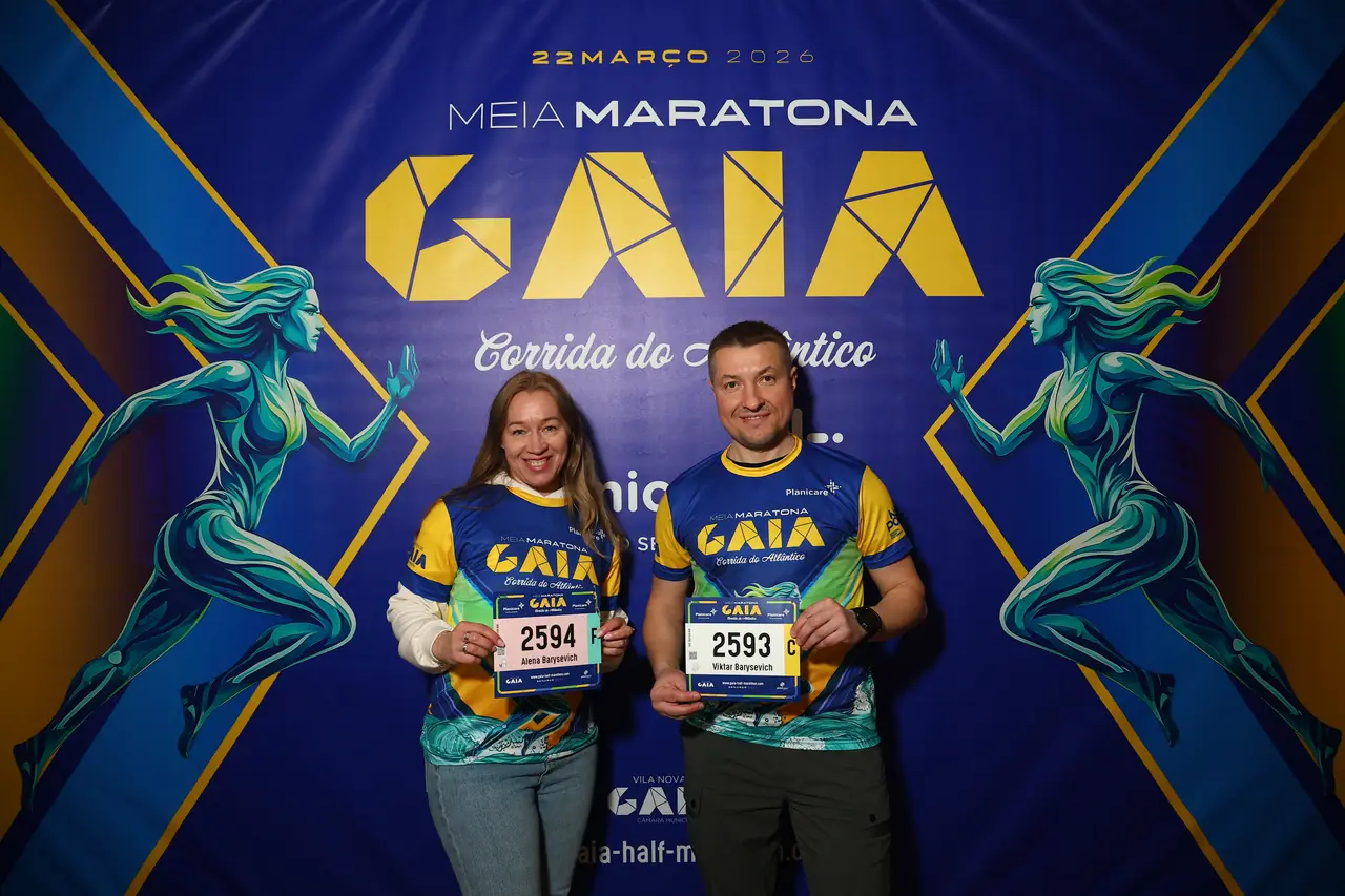 Dois participantes posam junto ao painel oficial da prova, com os dorsais na mão.