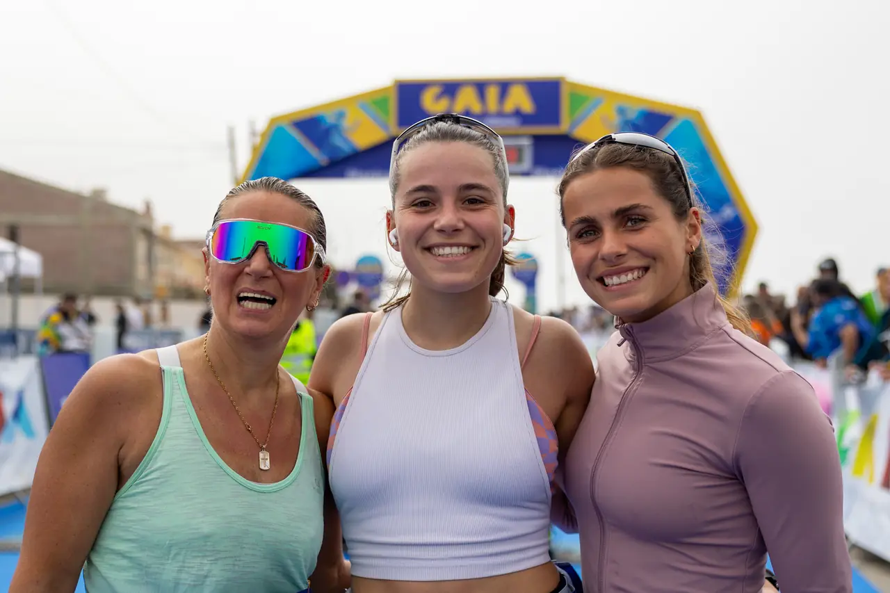 Três corredoras posam juntas junto ao pórtico da meta.