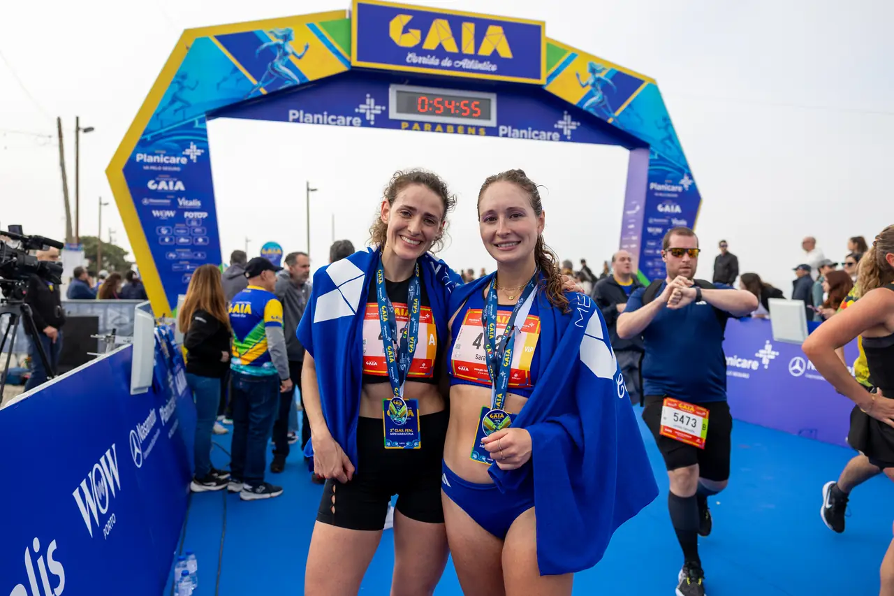 Duas corredoras com medalha ao pescoço, sob o pórtico da meta.