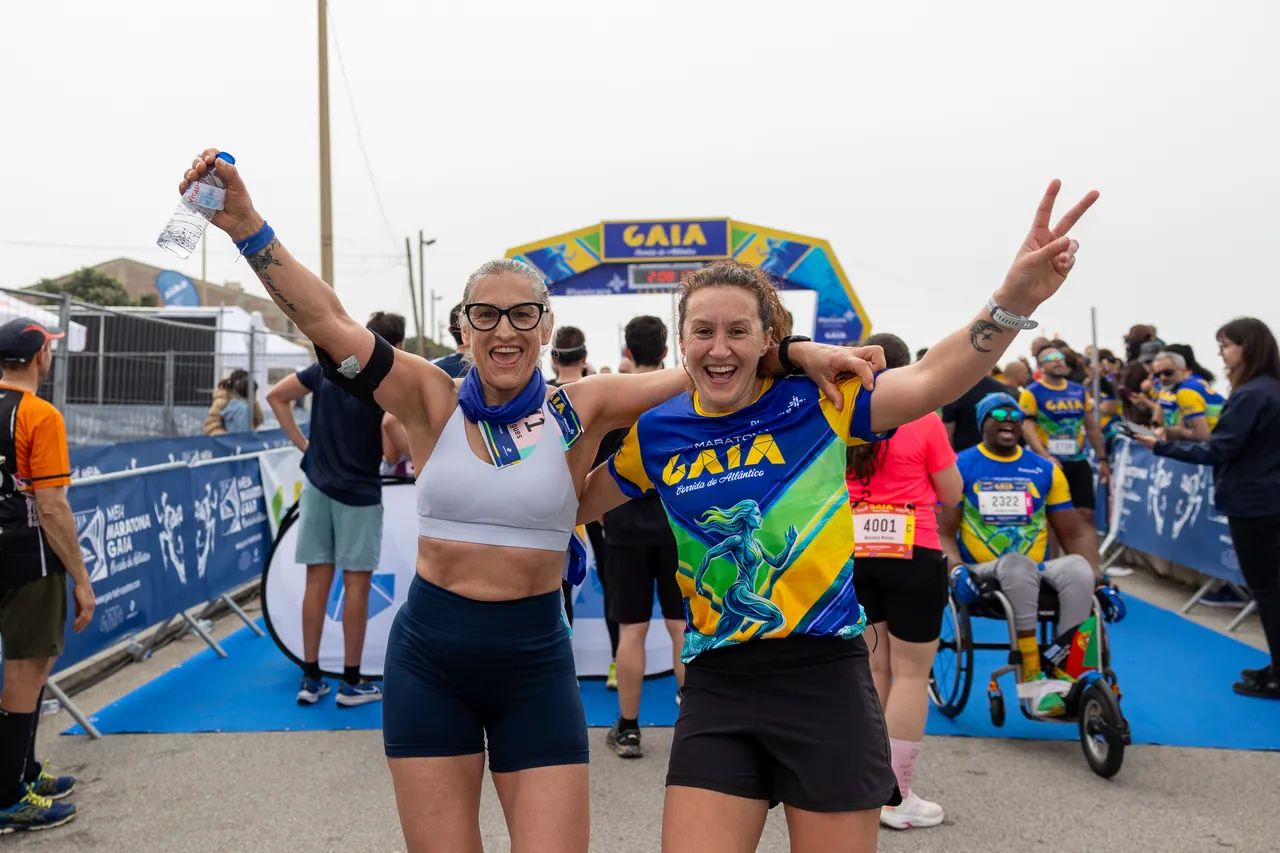 Duas corredoras erguem os braços e mostram as medalhas, a festejar juntas depois da chegada.