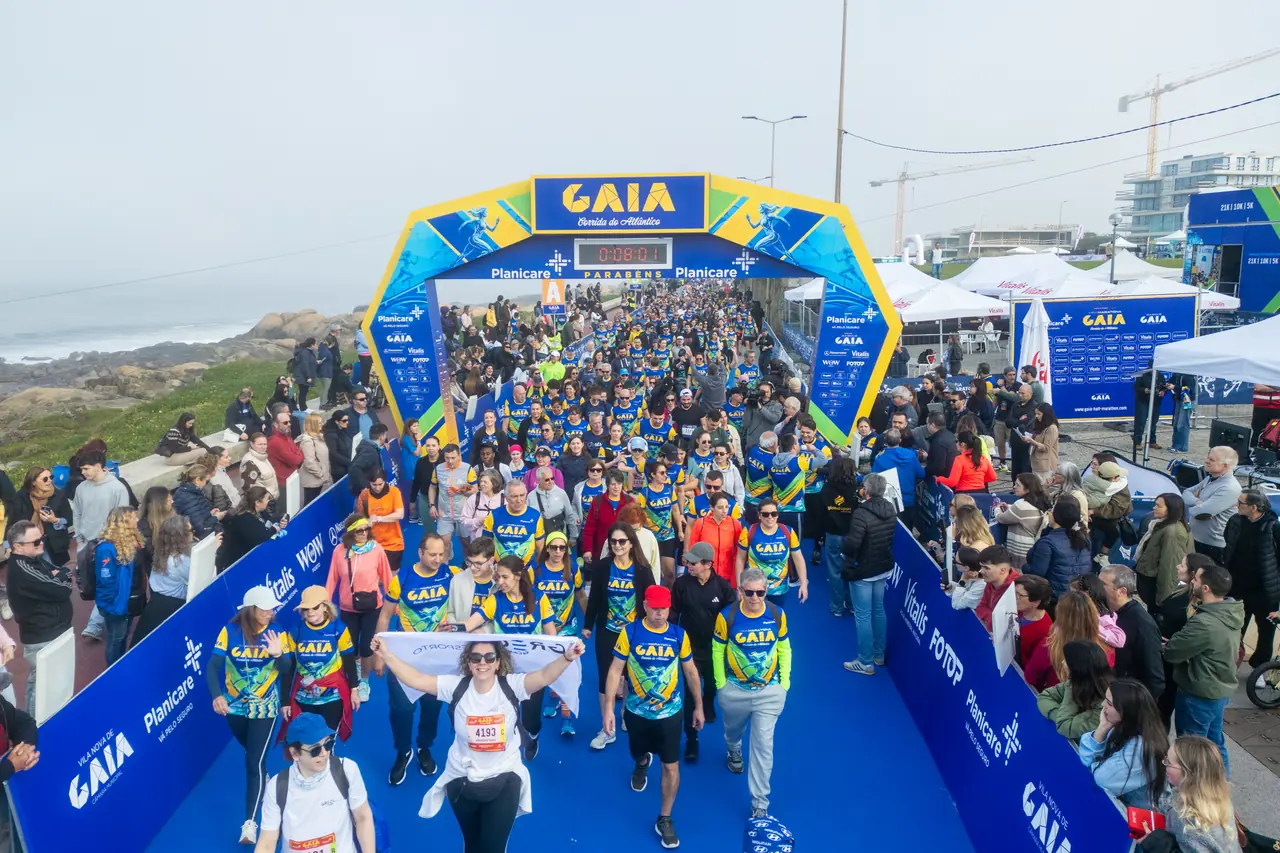 A vaga da caminhada solidária a passar sob o arco de partida em 2026: centenas de participantes de camisola da prova, o público encostado às grades dos dois lados e o oceano Atlântico à esquerda.