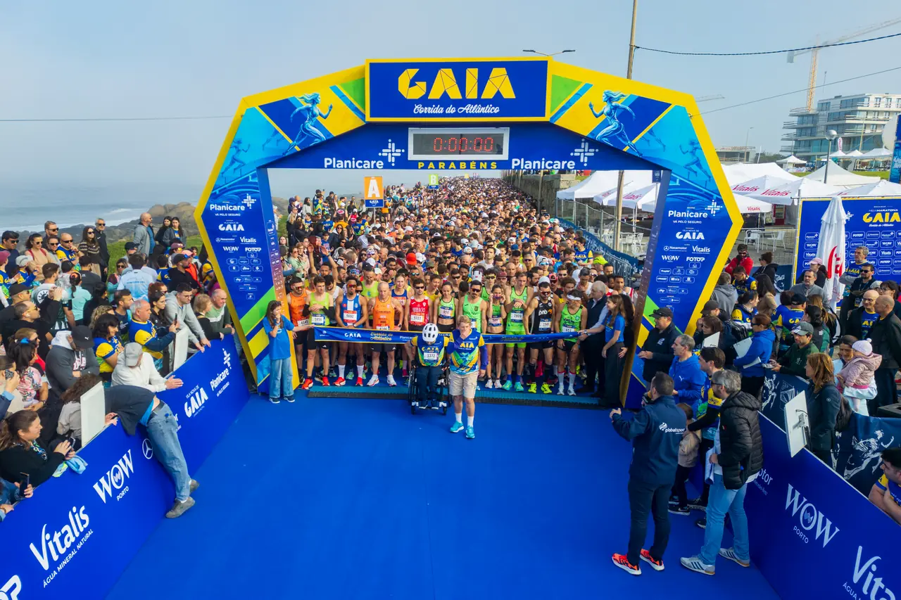 A largada dos 21 quilómetros de 2026: o pelotão de elite alinhado sob o arco com o relógio a zeros, um atleta em cadeira de rodas à frente e o público a encher as duas margens.